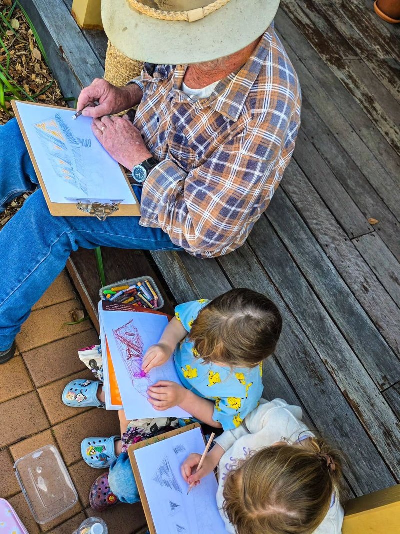 An older man and two young children drawing together on a porch