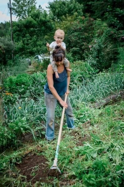 A mother hoeing a garden bed with her baby on her shoulders